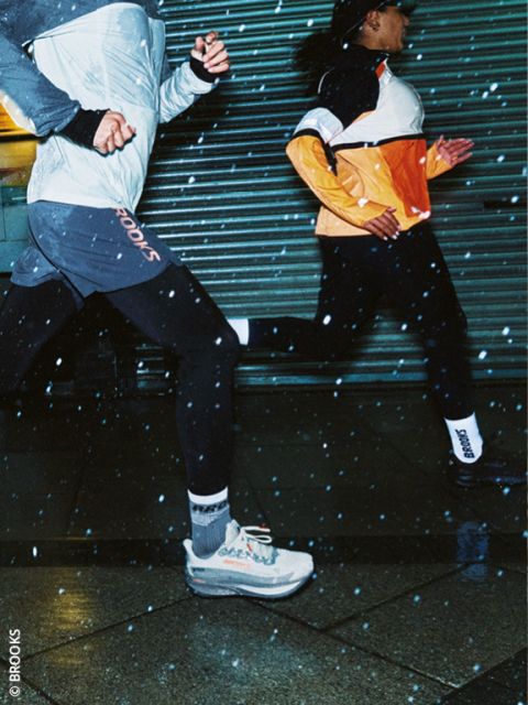 Zwei Personen, die nachts in Sportkleidung laufen, mit sichtbaren Regentropfen in der Luft und nassem Bürgersteig unter ihnen. Beide Läufer tragen Jacken und Laufschuhe und laufen schnell an einem Metalltor vorbei.