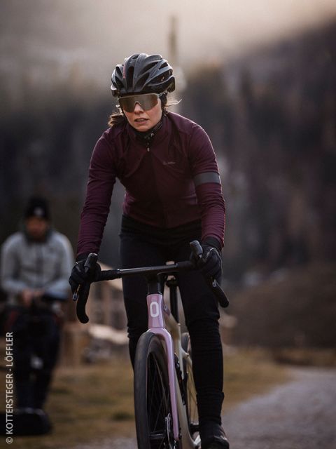 Ein Radfahrer mit Helm, Sonnenbrille, Handschuhen und kastanienbrauner Jacke fährt mit seinem Rennrad auf einem Schotterweg in einer bergigen Gegend. Der Hintergrund ist leicht unscharf, in der Ferne ist eine weitere Person zu sehen.