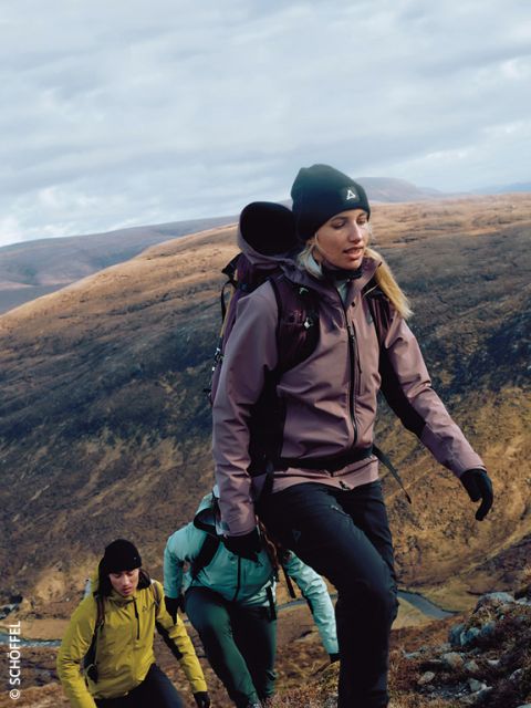 Drei Wanderer mit Outdoor-Ausrüstung und Rucksäcken erklimmen einen felsigen Hang mit braunen, sanften Hügeln im Hintergrund unter einem bewölkten Himmel. Der führende Wanderer trägt eine lila Jacke und eine Mütze.