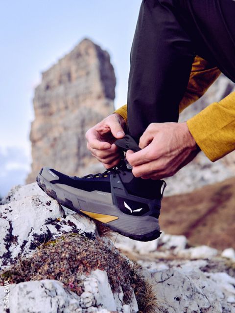 Eine Person in Outdoor-Kleidung zieht die Schnürsenkel eines schwarzen Wanderstiefels fest, während sie auf einer felsigen Oberfläche steht, mit einer großen Felsformation und blauem Himmel im Hintergrund.