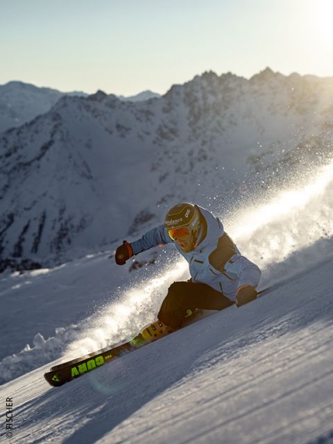Ein Skifahrer in blauer Jacke und mit Helm fährt bei Sonnenuntergang einen verschneiten Berghang hinunter und wirbelt dabei Pulverschnee auf, während im Hintergrund schroffe, sonnenbeschienene Berggipfel zu sehen sind.