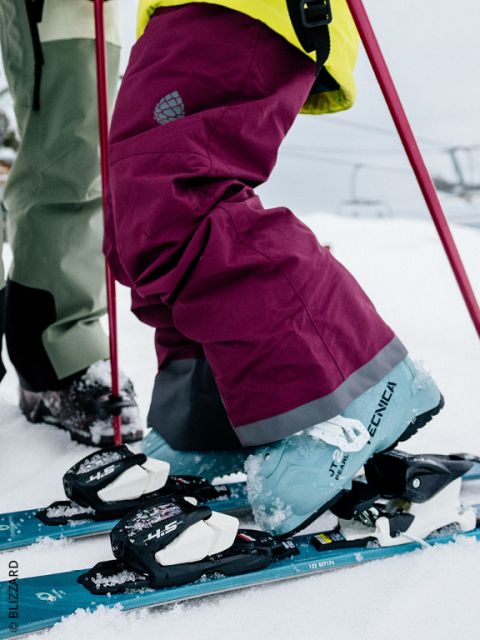 Nahaufnahme der Beine eines Skifahrers, der eine kastanienbraune Skihose und blaue Tecnica-Skischuhe trägt, die in die Bindungen im Schnee eingeklinkt sind, mit roten Skistöcken und einem Skilift im Hintergrund.