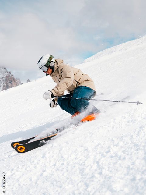 Ein Skifahrer in beiger Jacke, blauer Hose und weißem Helm fährt auf einer verschneiten Piste bergab, macht eine scharfe Kurve und wirbelt Schnee auf. Im Hintergrund sind Berge zu sehen.