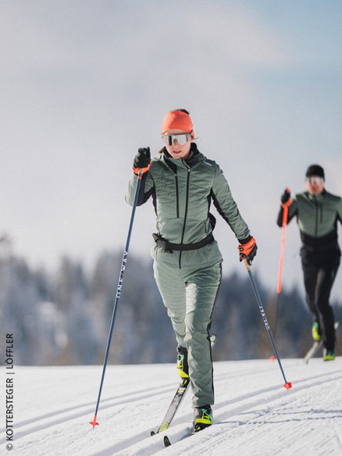 Zwei Skilangläufer auf einer verschneiten Loipe, mit Wintersportausrüstung und Sonnenbrille, mit Bäumen und blauem Himmel im Hintergrund.