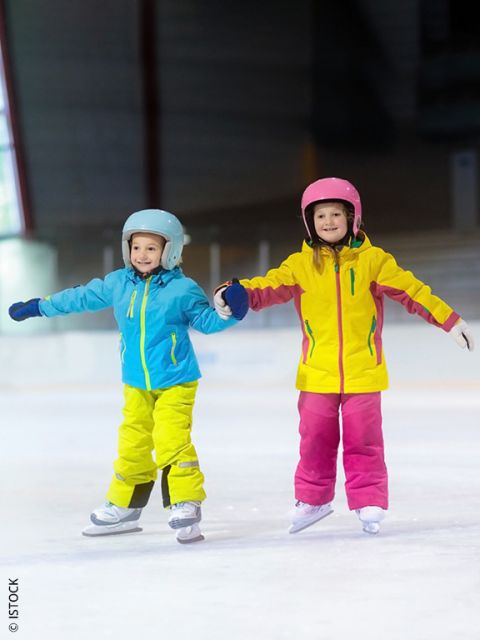 Zwei kleine Kinder in bunten Jacken und Helmen laufen gemeinsam Schlittschuh, halten sich an den Händen und lächeln auf einer Eislaufbahn.