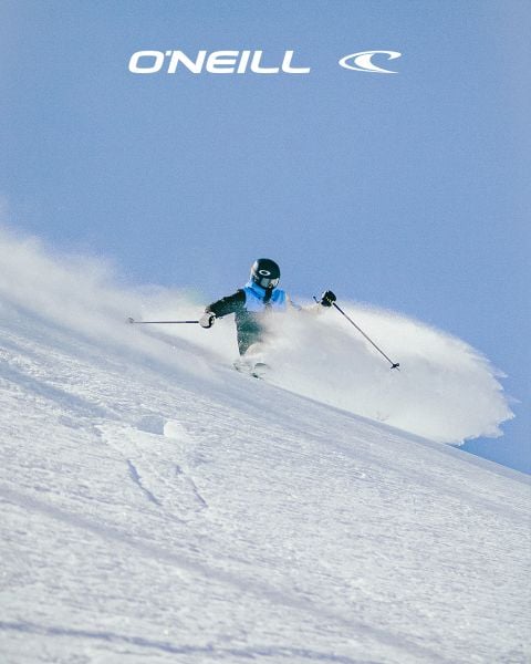 Eine Frau mit langen Haaren steht in einer Wüstenlandschaft unter einem blauen Himmel mit vereinzelten Wolken. Sie trägt ein schwarzes O'Neill-Tanktop und lehnt sich leicht nach vorne. Auf der rechten Seite des Bildes ist das O'Neill-Logo zu sehen.