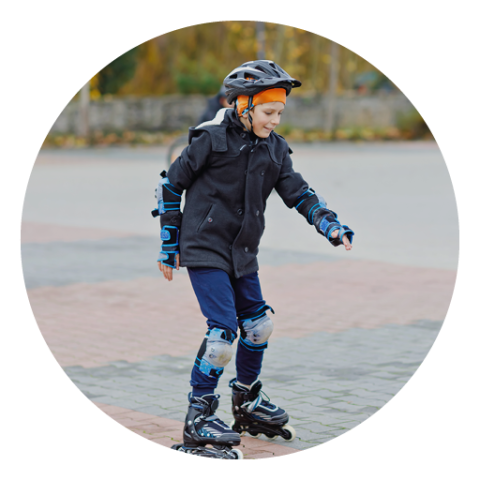 Ein Kind mit Helm, dunkler Jacke und Schonern fährt im Freien auf Rollschuhen auf einer gepflasterten Fläche. Im Hintergrund sind Bäume und eine Mauer zu sehen.