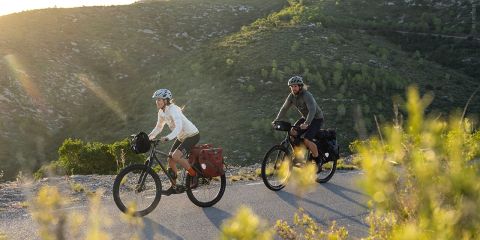 Zwei Radfahrer mit Helmen und Outdoor-Ausrüstung fahren mit beladenen Tourenrädern auf einer asphaltierten Straße durch eine hügelige, grüne Landschaft im Sonnenlicht. Sträucher und Vegetation säumen den Vorder- und Hintergrund.