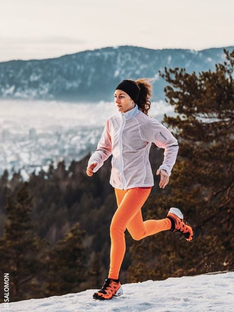 Eine Frau in einer weißen Jacke und leuchtend orangefarbenen Leggings läuft auf einem verschneiten Weg mit Kiefern und Bergen im Hintergrund. Der Himmel ist bewölkt und die Landschaft unter ihr ist teilweise mit Schnee bedeckt.