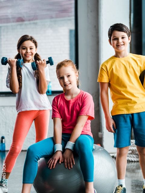 Drei lächelnde Kinder in Sportkleidung posieren in einer Turnhalle. Ein Mädchen hält Hanteln, ein anderes sitzt auf einem Gymnastikball, und ein Junge steht daneben. Sie wirken glücklich und bereit für körperliche Betätigung.
