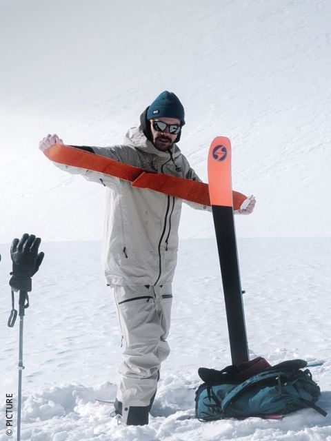 Eine Person in Winterausrüstung zieht auf einem verschneiten Berg ein orangefarbenes Steigfell über einen Ski, während sie Skistöcke und einen Rucksack bei sich trägt.