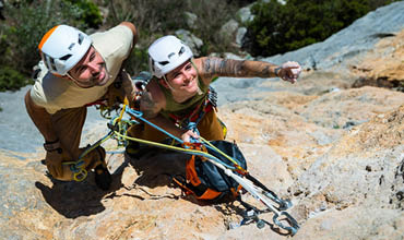 Zwei Kletterer mit Helmen und Ausrüstung machen an einem Felsen eine Pause und überprüfen gemeinsam im hellen Sonnenlicht ihre Seile und Ausrüstung.