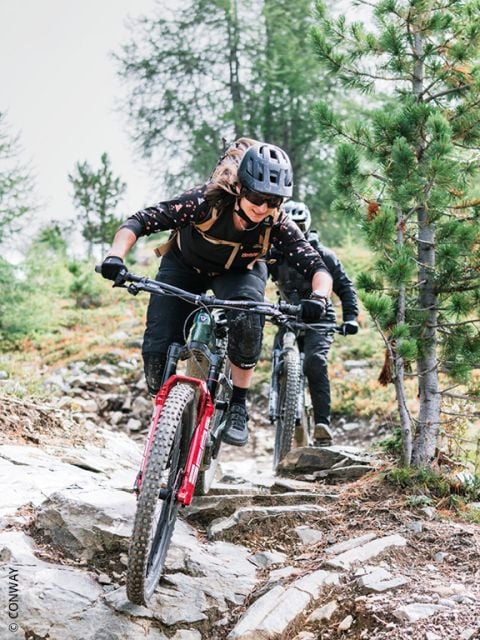 Zwei Radfahrer mit Helmen fahren mit ihren Mountainbikes einen felsigen, bewaldeten Weg hinunter, wobei sie sich konzentrieren und zwischen Bäumen und unebenem Gelände hindurchfahren.