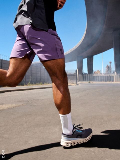 Person, die auf einer sonnenbeschienenen Straße unter einer Autobahnüberführung läuft, in lila Shorts, weißen Socken und grauen Laufschuhen.