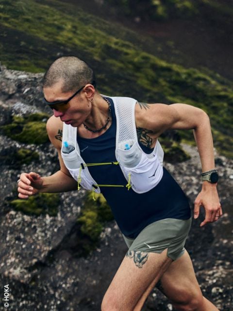 Eine Person mit Tattoos läuft auf einem steinigen, moosigen Weg bergauf, trägt eine Sonnenbrille, eine Trinksportweste und eine Smartwatch.