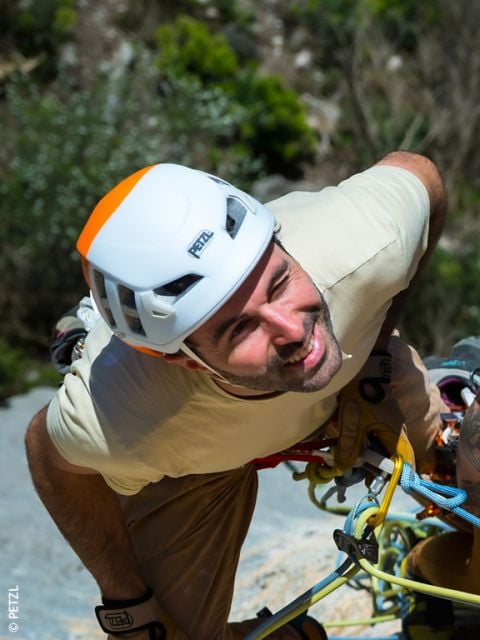 Ein Mann, der einen weißen Helm und eine Kletterausrüstung trägt, lächelt beim Klettern im Freien an einem sonnigen Tag.