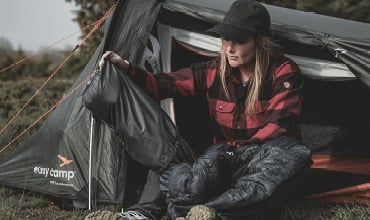 Eine Frau in einem rot karierten Hemd sitzt am Eingang eines Zeltes und zieht einen Schlafsack an oder passt ihn an, während sie im Freien zeltet.