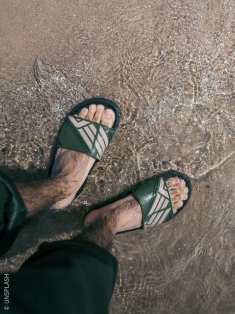 Eine Person mit grünen Sandalen steht im flachen Wasser an einem Sandstrand, die Hosenbeine leicht hochgekrempelt.