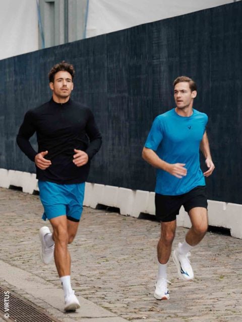Zwei Männer in Sportkleidung joggen auf einem Kopfsteinpflasterweg neben einem dunklen Zaun; beide tragen blaue Shorts und weiße Turnschuhe.