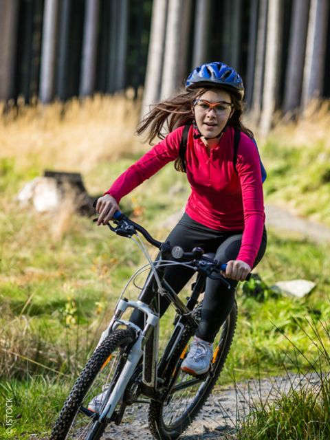 Eine Frau in einem roten Shirt und mit Helm fährt auf einem Waldweg im Sonnenlicht mit dem Mountainbike.