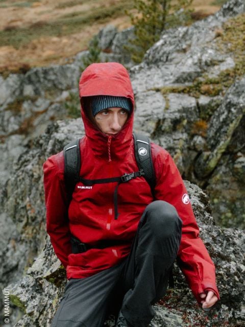 Eine Person in einer roten Kapuzenjacke und mit einem Rucksack sitzt draußen auf Felsen, im Hintergrund sind Bäume und felsiges Gelände zu sehen.