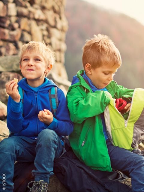 Zwei kleine Jungen sitzen draußen auf Felsen. Der eine isst einen Snack und lächelt, während der andere in einem grünen Rucksack herumwühlt. Beide tragen bunte Jacken.