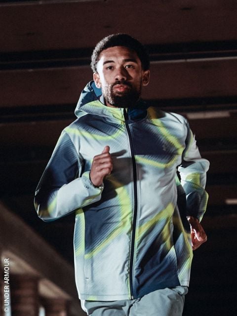 Eine Person, die eine Kapuzenjacke und Laufschuhe trägt, joggt an einem kalten, klaren Tag einen verschneiten Weg entlang, mit der Skyline der Stadt im Hintergrund.