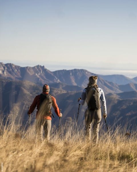 Zwei Wanderer mit Rucksäcken und Wanderstöcken stehen im hohen Gras und blicken unter klarem Himmel auf ferne Bergketten.